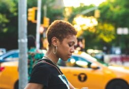 A Black woman with short curly hair looks down at her phone. There is a bright yellow taxi in the background.